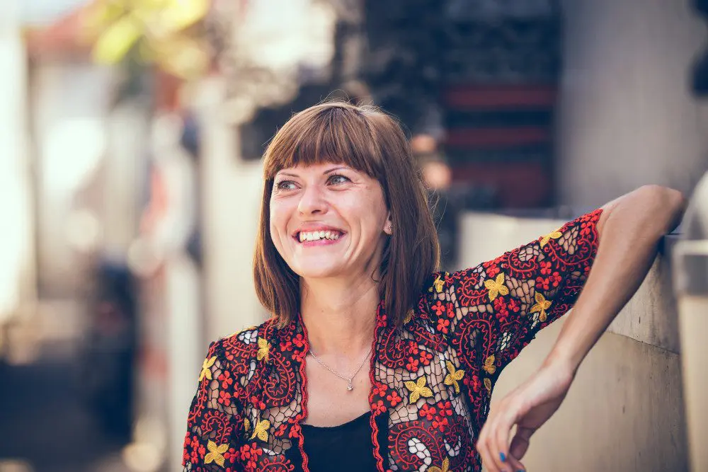 Woman with bangs and bob smiling while propping her elbow up against a concrete wall.