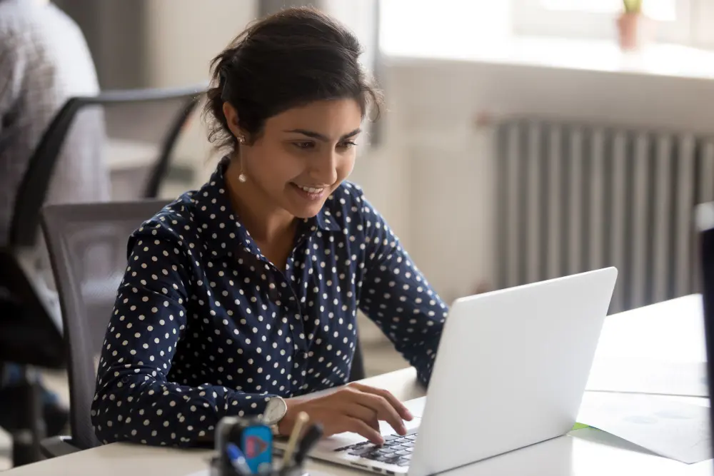 Smiling female employee using laptop at workplace, looking at screen, focused businesswoman doing computer work.
