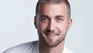 Portrait of a smiling man with short brown hair and light stubble, wearing a light gray shirt, against a pale blue background on a plain backdrop.